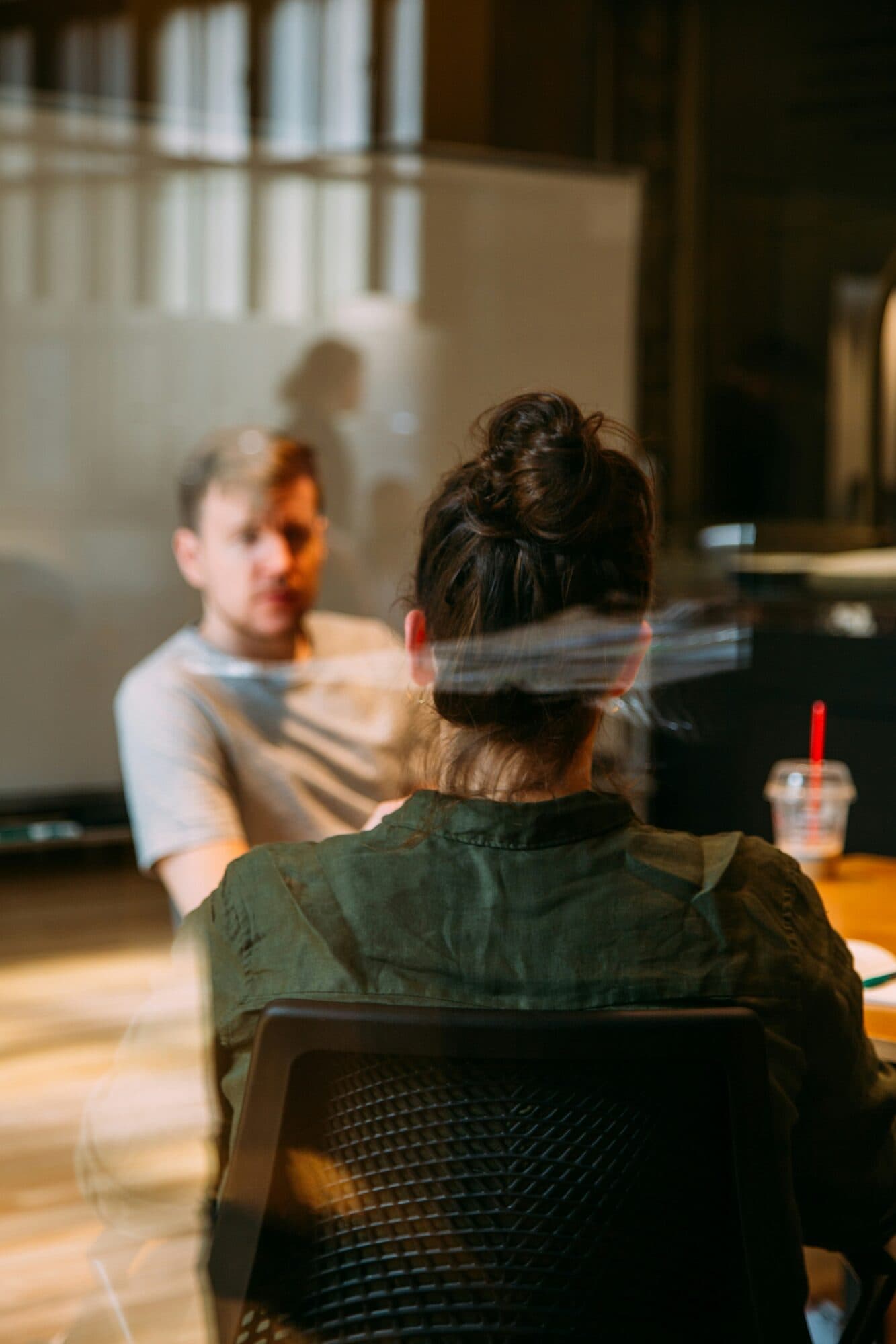 Two people in conversation across a table, seen through glass.